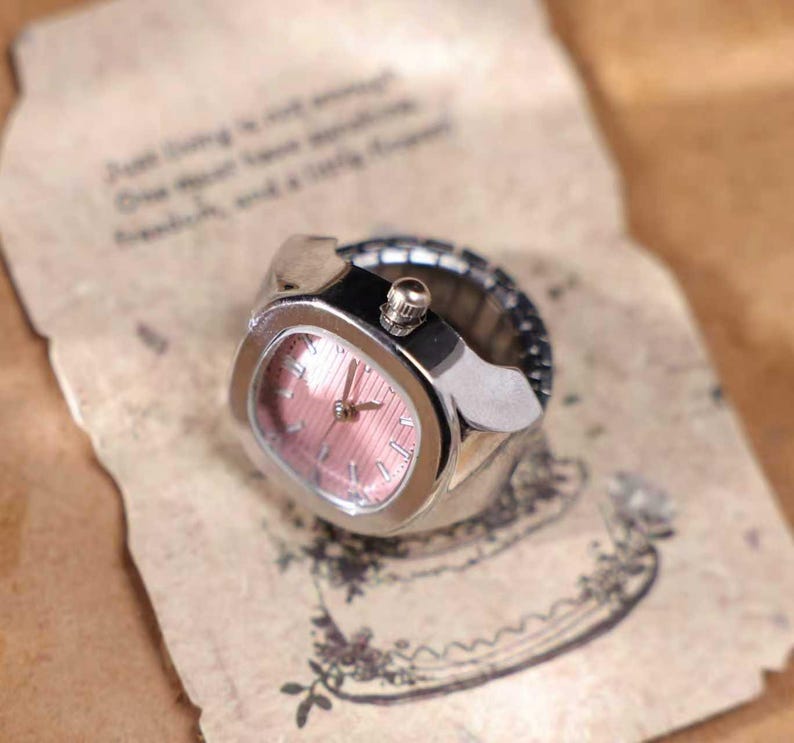 Puede incluir: Un reloj de anillo plateado con esfera rosa. El reloj tiene una esfera rectangular con una peque&ntilde;a corona. La correa es el&aacute;stica. El reloj est&aacute; sobre un papel con texto.