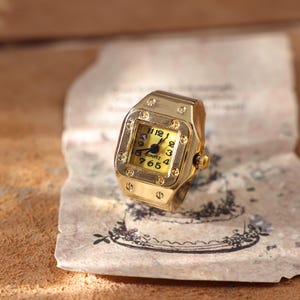 May include: A gold-toned watch ring with a square face and a yellow dial. The ring features a quartz movement and displays the numbers 1, 2, 3, 4, 5, 6, 7, 8, 9, 10, 11, and 12. The ring is resting on a piece of aged paper.