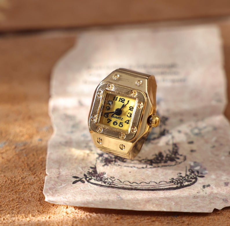 May include: A gold-toned watch ring with a square face and a yellow dial. The ring features a quartz movement and displays the numbers 1, 2, 3, 4, 5, 6, 7, 8, 9, 10, 11, and 12. The ring is resting on a piece of aged paper.