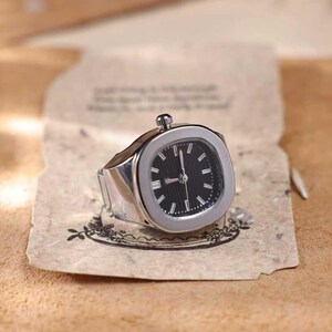 Puede incluir: Un reloj de anillo plateado con esfera negra e indicadores horarios blancos. El reloj tiene una esfera cuadrada con esquinas redondeadas y una peque&ntilde;a corona. El anillo est&aacute; sobre un papel envejecido.