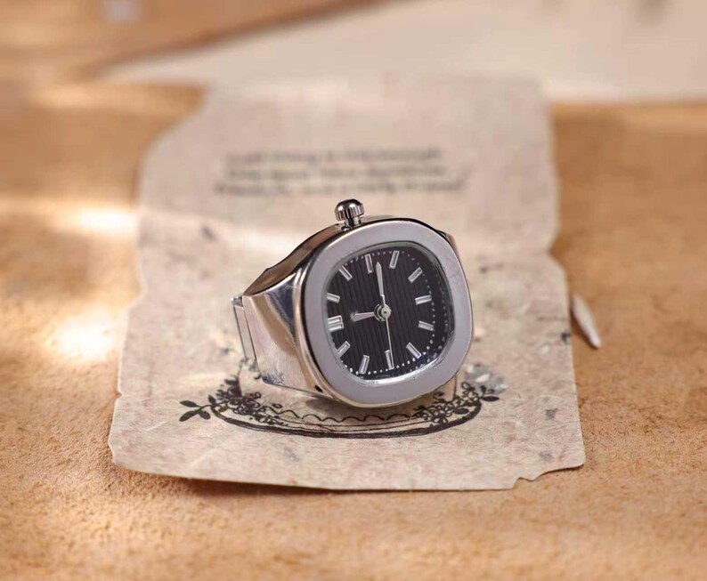 Puede incluir: Un reloj de anillo plateado con esfera negra e indicadores horarios blancos. El reloj tiene una esfera cuadrada con esquinas redondeadas y una peque&ntilde;a corona. El anillo est&aacute; sobre un papel envejecido.