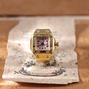 May include: A gold-toned quartz ring watch with a square face and a pink dial. The watch has black hands and numerals, and a small crown on the side. The ring is set on a piece of aged paper with a floral design.