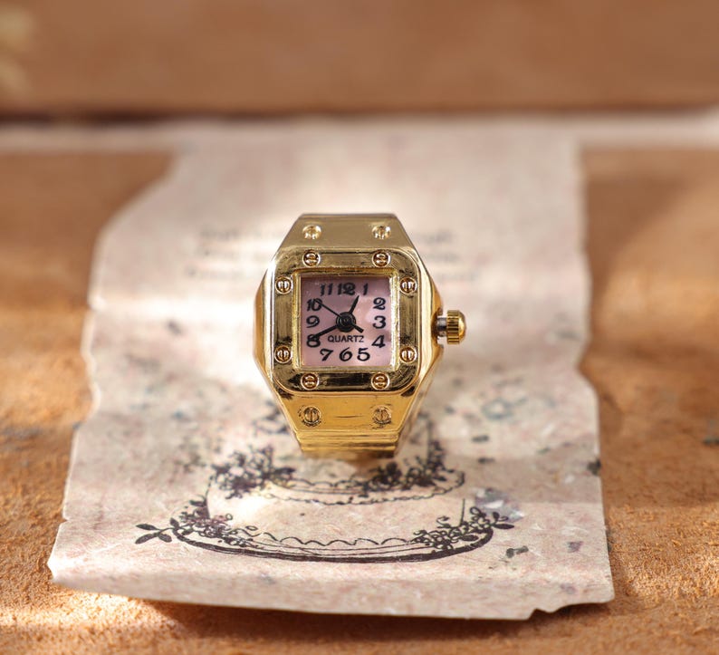 May include: A gold-toned quartz ring watch with a square face and a pink dial. The watch has black hands and numerals, and a small crown on the side. The ring is set on a piece of aged paper with a floral design.