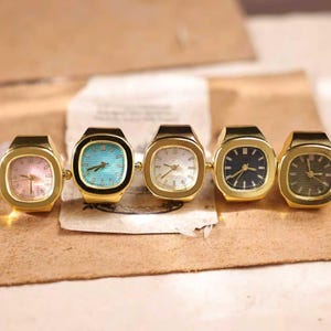 Puede incluir: Cinco anillos-reloj dorados con esferas de diferentes colores: rosa, turquesa, blanco, azul marino y verde oliva. Los relojes tienen forma rectangular con esquinas redondeadas y una pequeña corona en el lateral. Los relojes se muestran sobre una superficie marrón.