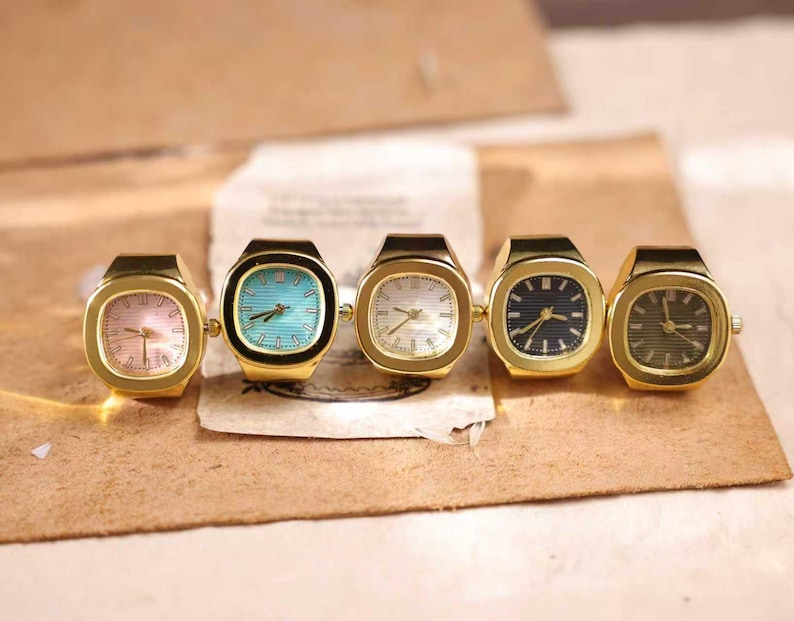 May include: Five gold-toned ring watches with different colored faces: pink, turquoise, white, navy, and olive green. The watches have a rectangular shape with rounded corners and a small crown on the side. The watches are displayed on a brown surface.