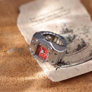May include: A silver-toned ring watch with a red face. The watch has a square face with silver screws and a stretch band. The watch is resting on a piece of aged paper with faded ink.