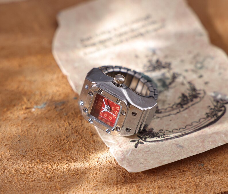 May include: A silver-toned ring watch with a red face. The watch has a square face with silver screws and a stretch band. The watch is resting on a piece of aged paper with faded ink.