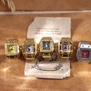 May include: Five square-faced ring watches in gold and silver tones. The watches have different colored faces, including yellow, white, black, pink, and red. The rings are displayed on a brown surface with a handwritten note.