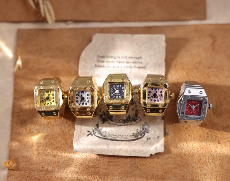 May include: Five square-faced ring watches in gold and silver tones. The watches have different colored faces, including yellow, white, black, pink, and red. The rings are displayed on a brown surface with a handwritten note.