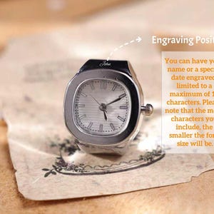 Puede incluir: Un reloj de anillo plateado con una esfera rectangular y una esfera de color claro. El reloj tiene una peque&ntilde;a corona y la palabra "John" est&aacute; grabada en la parte superior. La imagen incluye texto sobre grabar un nombre o fecha.