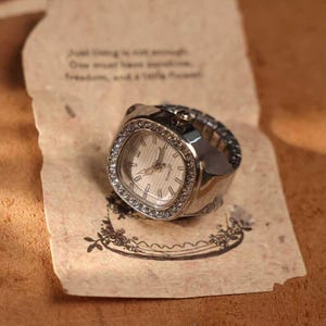 May include: A silver-toned ring watch with a square face and a white dial. The watch is adorned with small, clear crystals around the face. The watch rests on a piece of paper with handwritten text and a floral design.