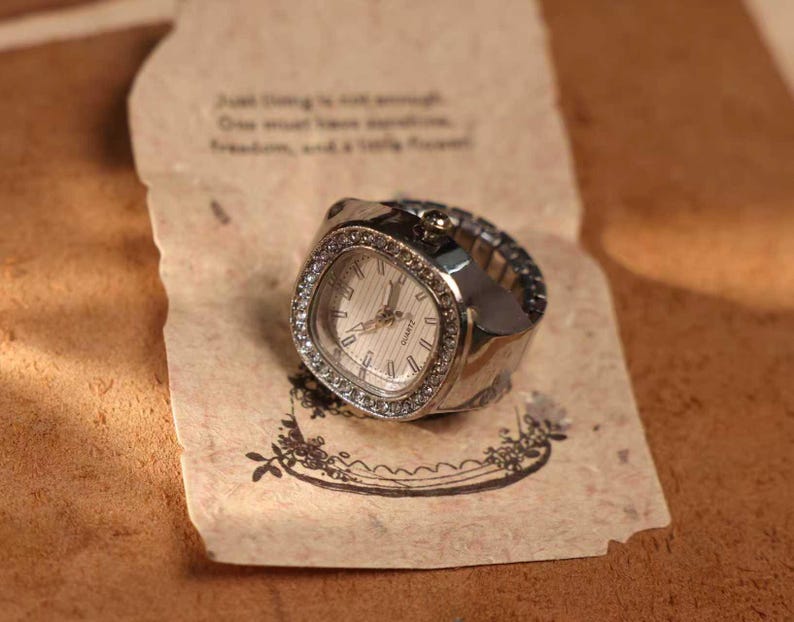 May include: A silver-toned ring watch with a square face and a white dial. The watch is adorned with small, clear crystals around the face. The watch rests on a piece of paper with handwritten text and a floral design.