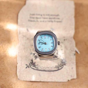 Puede incluir: Un reloj de anillo plateado con esfera azul claro y n&uacute;meros romanos. El reloj tiene una esfera rectangular y una peque&ntilde;a corona. El anillo est&aacute; sobre un fondo de papel vintage con texto manuscrito.