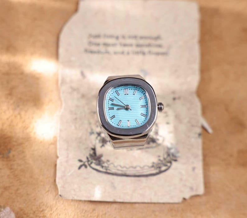 Puede incluir: Un reloj de anillo plateado con esfera azul claro y n&uacute;meros romanos. El reloj tiene una esfera rectangular y una peque&ntilde;a corona. El anillo est&aacute; sobre un fondo de papel vintage con texto manuscrito.