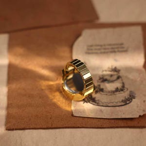 May include: A gold-toned ring with a segmented band and a dark, reflective center. The ring sits on a textured, brown surface next to a vintage paper with a cake illustration.