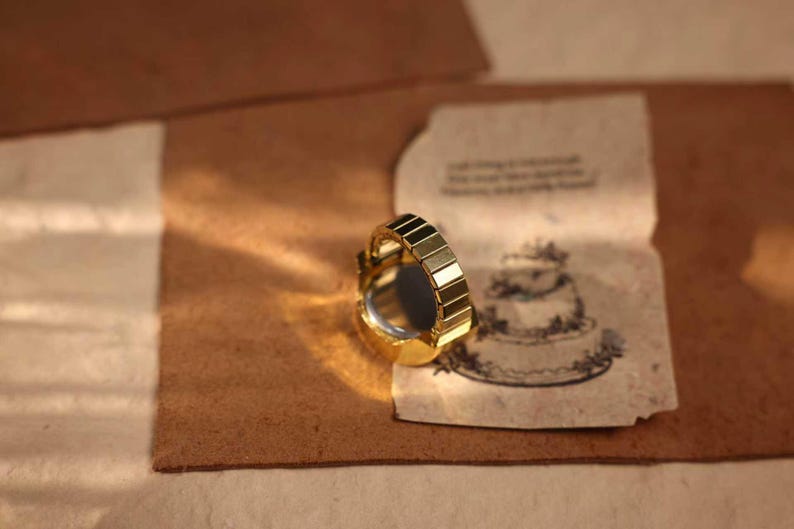 May include: A gold-toned ring with a segmented band and a dark, reflective center. The ring sits on a textured, brown surface next to a vintage paper with a cake illustration.