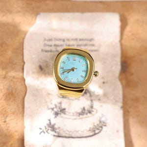 May include: A gold-toned ring watch with a square face and a light blue dial. The watch has Roman numeral hour markers and a gold band. The ring is set on a vintage paper with text and a floral design.