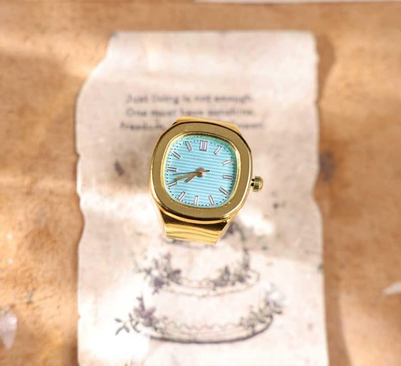 May include: A gold-toned ring watch with a square face and a light blue dial. The watch has Roman numeral hour markers and a gold band. The ring is set on a vintage paper with text and a floral design.