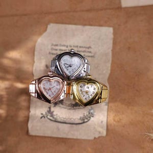 May include: Three heart-shaped ring watches in silver, rose gold, and gold tones. Each watch has a white face with black numerals and hands. The rings are displayed on a vintage paper with handwritten text.