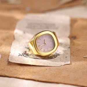 May include: A gold-colored ring with a watch face. The watch face is a light pink color with gold-colored hands and hour markers. The ring is sitting on a piece of aged paper.