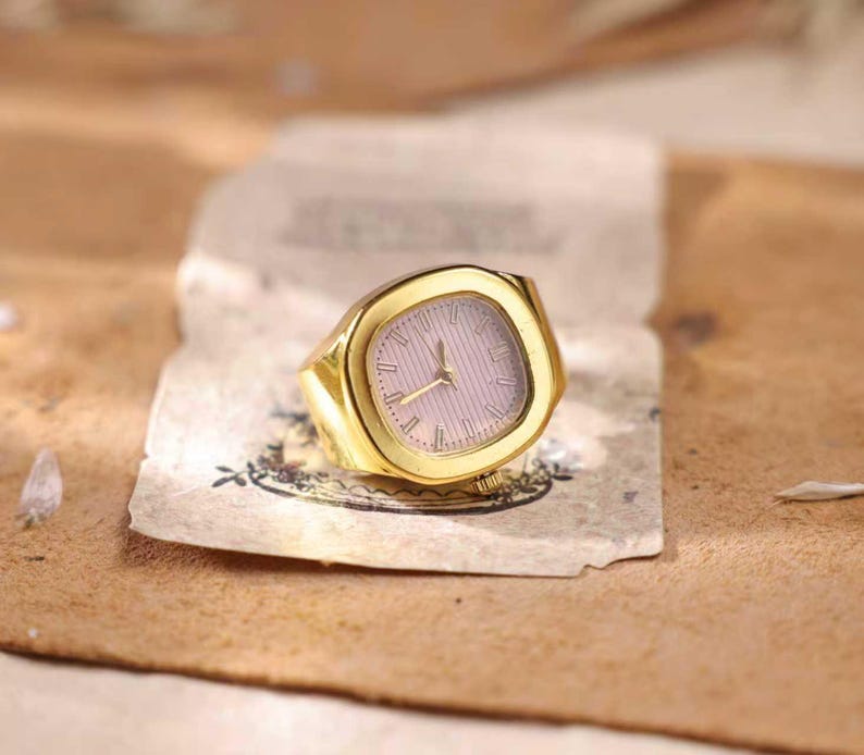 May include: A gold-colored ring with a watch face. The watch face is a light pink color with gold-colored hands and hour markers. The ring is sitting on a piece of aged paper.