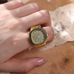 May include: A gold-toned ring watch with a rectangular face and a light green dial. The watch has a classic design with hour markers and hands. The ring is worn on a finger.