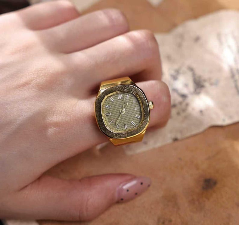 May include: A gold-toned ring watch with a rectangular face and a light green dial. The watch has a classic design with hour markers and hands. The ring is worn on a finger.