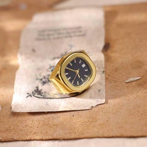 May include: A gold-toned ring with a watch face. The watch face is black with gold hands and markers. The ring has a square face with rounded corners and a thick band. The ring is set on a piece of aged paper.