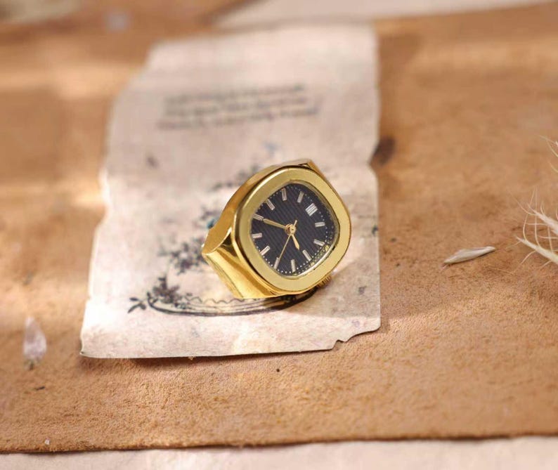 May include: A gold-toned ring with a watch face. The watch face is black with gold hands and markers. The ring has a square face with rounded corners and a thick band. The ring is set on a piece of aged paper.
