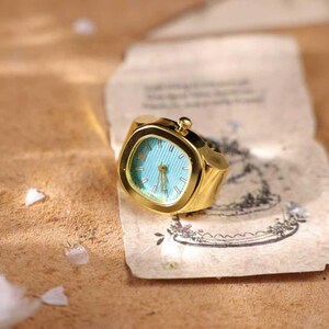 May include: A gold-toned ring watch with a square face and a light blue dial. The watch has a small crown and is set on a piece of aged paper with a decorative illustration. The ring is a unique piece of jewelry.