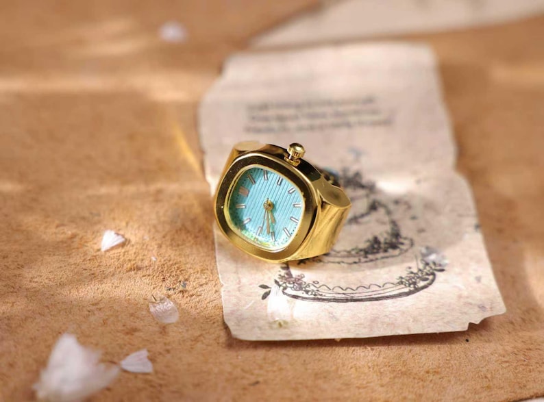 May include: A gold-toned ring watch with a square face and a light blue dial. The watch has a small crown and is set on a piece of aged paper with a decorative illustration. The ring is a unique piece of jewelry.