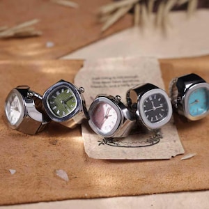 Puede incluir: Cinco anillos-reloj plateados con esferas cuadradas. Las esferas muestran diferentes colores, incluyendo blanco, verde, rosa, negro y turquesa. Los anillos se exhiben sobre una superficie marr&oacute;n.