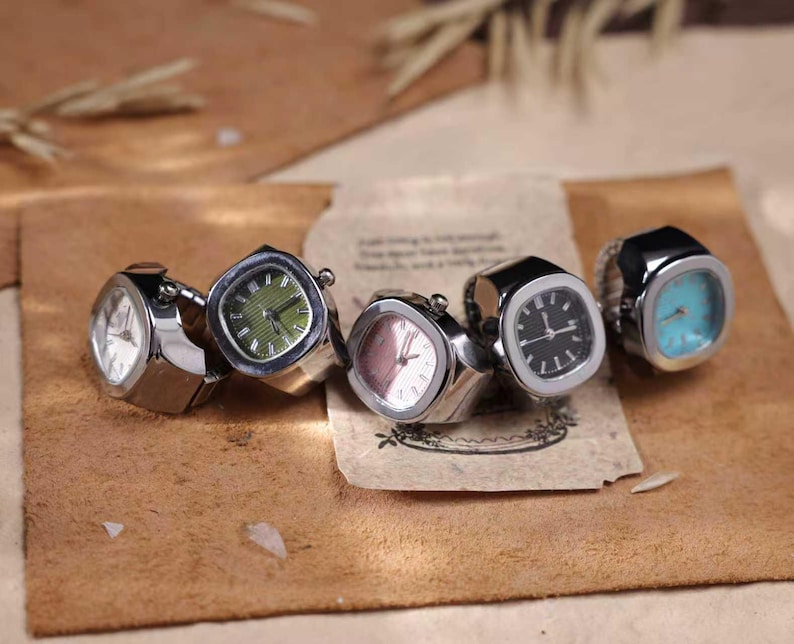 Puede incluir: Cinco anillos-reloj plateados con esferas cuadradas. Las esferas muestran diferentes colores, incluyendo blanco, verde, rosa, negro y turquesa. Los anillos se exhiben sobre una superficie marr&oacute;n.
