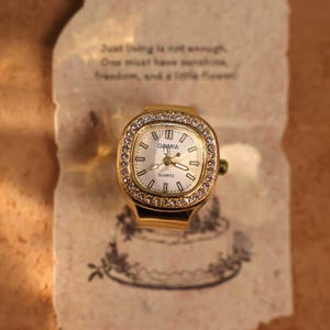 May include: A gold-toned ring watch with a rectangular face, surrounded by small, clear crystals. The watch face is white with black numerals and hands, and the brand name "GAMIA" is visible. The watch rests on a piece of paper with text.