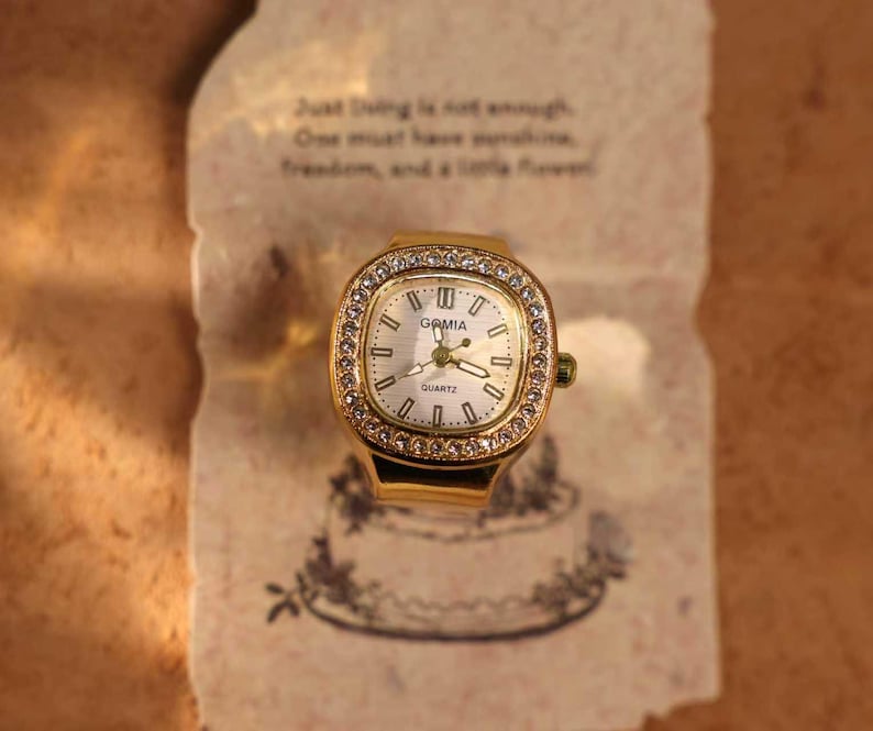 May include: A gold-toned ring watch with a rectangular face, surrounded by small, clear crystals. The watch face is white with black numerals and hands, and the brand name "GAMIA" is visible. The watch rests on a piece of paper with text.