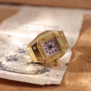 May include: A gold-toned quartz watch ring with a square face and a pink dial. The watch ring has a gold band and a small crown. The watch face displays black numerals and hands. The watch is resting on a piece of paper.
