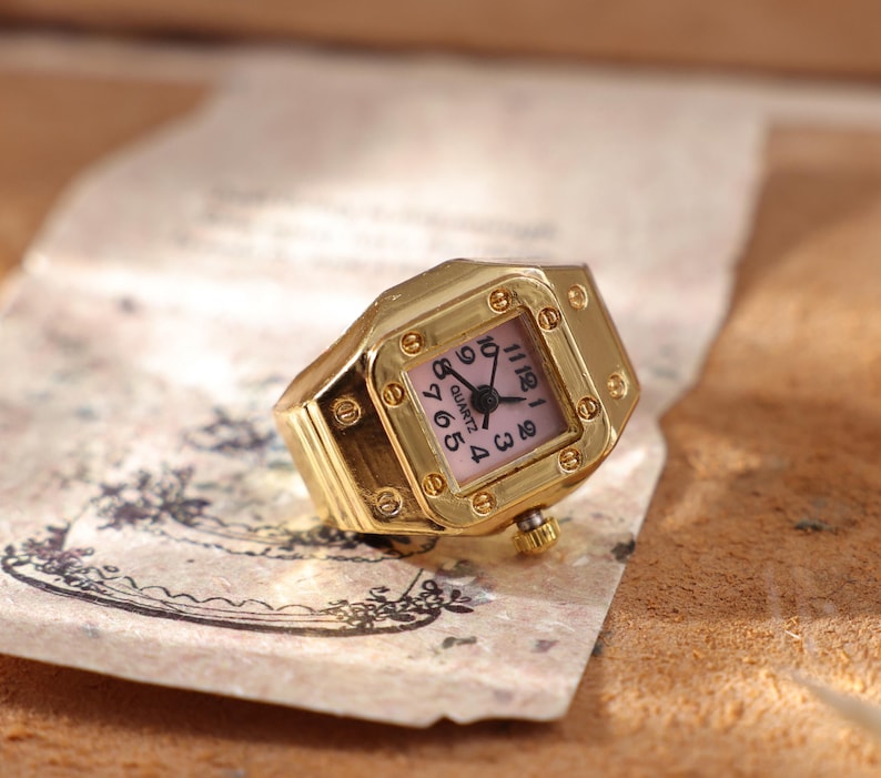 May include: A gold-toned quartz watch ring with a square face and a pink dial. The watch ring has a gold band and a small crown. The watch face displays black numerals and hands. The watch is resting on a piece of paper.