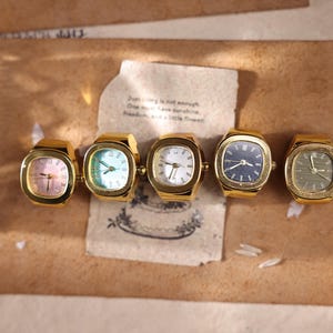 May include: Five vintage gold-toned ring watches with square faces. The watch faces display various colors, including pink, blue, white, and green. The watches are arranged on a brown surface with a handwritten note.