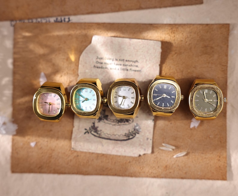 May include: Five vintage gold-toned ring watches with square faces. The watch faces display various colors, including pink, blue, white, and green. The watches are arranged on a brown surface with a handwritten note.