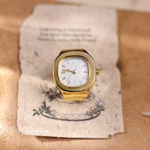 May include: A gold-toned ring watch with a white face and gold hands. The watch has a rectangular face with a gold bezel. The watch is set on a piece of aged paper with floral illustrations.
