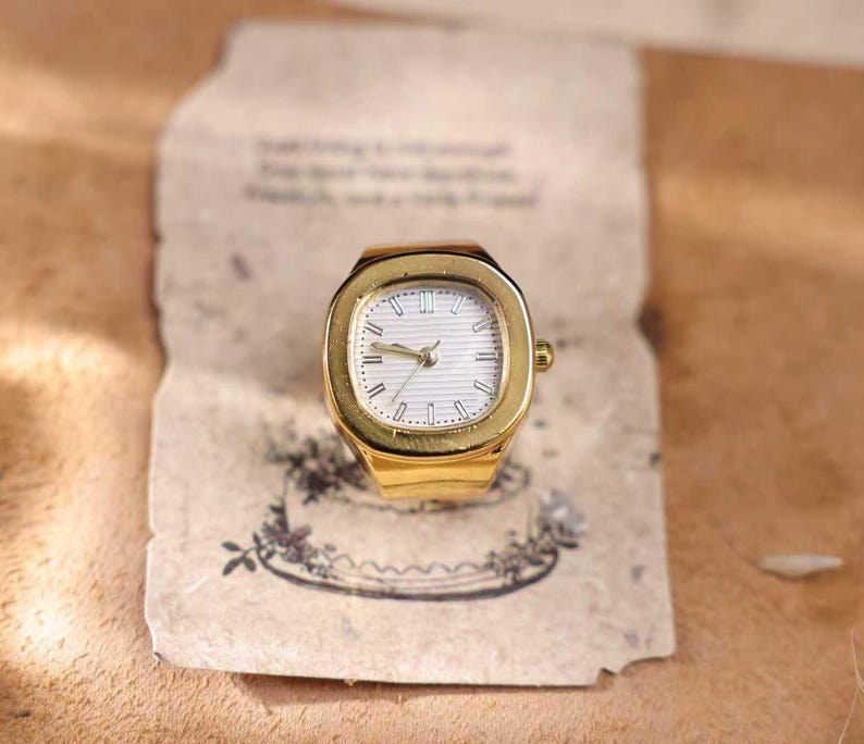 May include: A gold-toned ring watch with a white face and gold hands. The watch has a rectangular face with a gold bezel. The watch is set on a piece of aged paper with floral illustrations.
