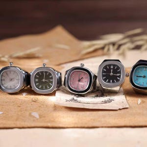 Può includere: Cinque anelli color argento con quadranti di orologio in vari colori: bianco, verde, rosa, nero e turchese. I quadranti sono quadrati o rettangolari. Gli anelli sono esposti su una superficie marrone.