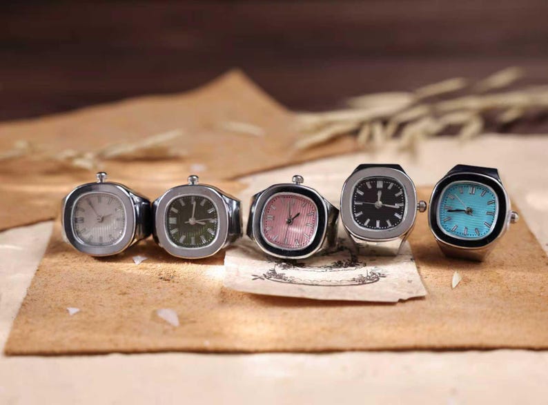 Puede incluir: Cinco anillos plateados con esferas de reloj en varios colores: blanco, verde, rosa, negro y turquesa. Las esferas son cuadradas o rectangulares. Los anillos se exhiben sobre una superficie marr&oacute;n.