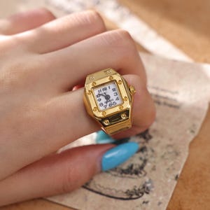 May include: A gold-colored ring shaped like a wristwatch. The ring has a square face with a white background and black numbers and hands. The ring is worn on a finger.