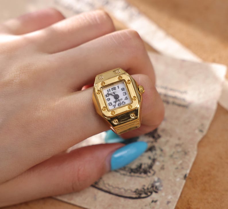 May include: A gold-colored ring shaped like a wristwatch. The ring has a square face with a white background and black numbers and hands. The ring is worn on a finger.