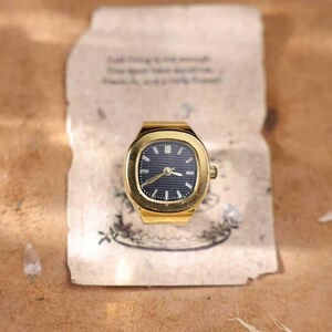 May include: A gold-toned watch ring with a dark blue face and gold hands. The watch has a rectangular face with rounded corners. The watch is set on a piece of paper with faded text and a floral design.