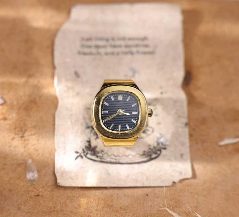May include: A gold-toned watch ring with a dark blue face and gold hands. The watch has a rectangular face with rounded corners. The watch is set on a piece of paper with faded text and a floral design.