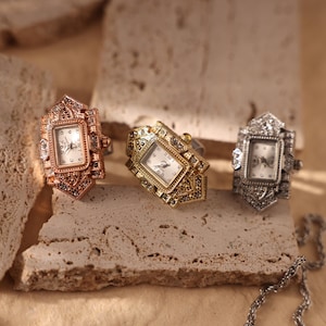 Puede incluir: Tres relojes de anillo ornamentados en oro rosa, oro y plata. Cada reloj tiene una esfera rectangular con una esfera blanca y números negros. Los anillos están decorados con patrones intrincados y pequeñas piedras preciosas, colocados sobre una superficie texturizada.