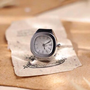 Puede incluir: Un reloj de anillo plateado con una esfera cuadrada y una esfera de color claro. El reloj tiene una peque&ntilde;a corona y est&aacute; colocado sobre un trozo de papel envejecido con un dise&ntilde;o floral. Las manecillas del reloj se&ntilde;alan las 10:10.