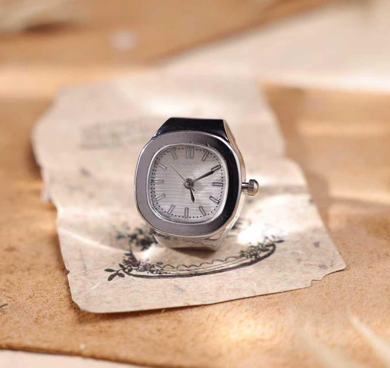 Puede incluir: Un reloj de anillo plateado con una esfera cuadrada y una esfera de color claro. El reloj tiene una peque&ntilde;a corona y est&aacute; colocado sobre un trozo de papel envejecido con un dise&ntilde;o floral. Las manecillas del reloj se&ntilde;alan las 10:10.